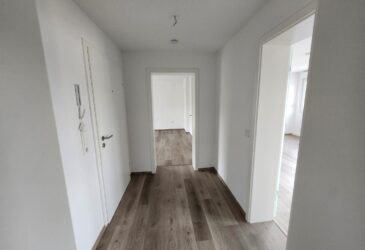 Das Bild zeigt die Diele der 3-Zimmer-Wohnung in Iserlohn-Letmathe. Wände und Türen der Diele sind weiß gestrichen. Rechts neben der Eingangstüre befindet sich die Bedienung der neuen Haussprechanlage. Die Diele verfügt über Tageslicht, da das Wohnzimmer ein Glastüre hat. Der Fußboden ist mit Vinyl in grau-braunem Holz Dekor ausgelegt.