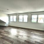 Wohnzimmer mit drei zweiflügeligen Fenstern nach Westen gerichtet. Links neben den Fenstern befindet sich ein Heizkörper. Rechts neben den Fenstern ist ebenfalls ein Heizkörper. Der Fußboden ist Vinyl in grau-braunem Holzdekor ausgelegt.