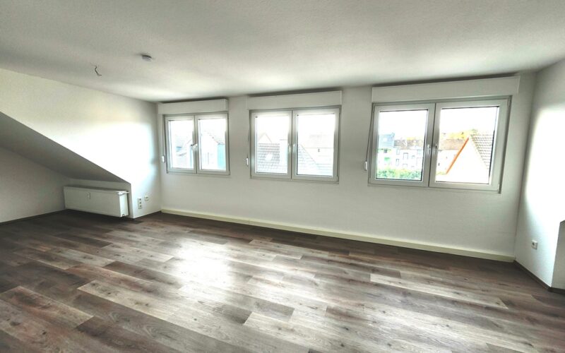 Wohnzimmer mit drei zweiflügeligen Fenstern nach Westen gerichtet. Links neben den Fenstern befindet sich ein Heizkörper. Rechts neben den Fenstern ist ebenfalls ein Heizkörper. Der Fußboden ist Vinyl in grau-braunem Holzdekor ausgelegt.
