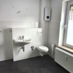 Waschtisch und WC im Bad sind mit einer Vorwand-Installation montiert. Darüber ist der Durchlauferhitzer für Küche und Bad, Die Wände sich weiß gestrichen und im Spritzbereich weiß gefliest. der Fußboden ist anthrazit gefliest. Zwischen Bad und WC befindet sich der Wasserzähler.