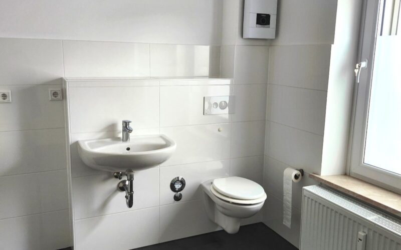 Waschtisch und WC im Bad sind mit einer Vorwand-Installation montiert. Darüber ist der Durchlauferhitzer für Küche und Bad, Die Wände sich weiß gestrichen und im Spritzbereich weiß gefliest. der Fußboden ist anthrazit gefliest. Zwischen Bad und WC befindet sich der Wasserzähler.