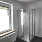 Das Bad mit Eck-Dusche. Neben der Dusche befindet sich ein großes Fenster. Unter dem Fenster ist ein Heizkörper