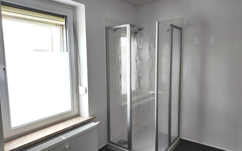 Das Bad mit Eck-Dusche. Neben der Dusche befindet sich ein großes Fenster. Unter dem Fenster ist ein Heizkörper