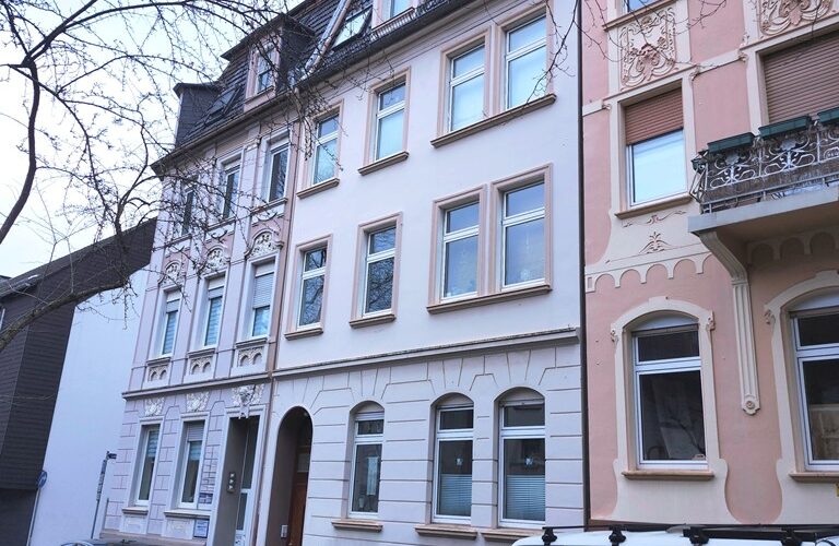 Häuserfronten mit dem Mehrfamilienhaus in Iserlohn. Vor dem Haus parken Autos. Die Straße ist verkehrsberuhigt.