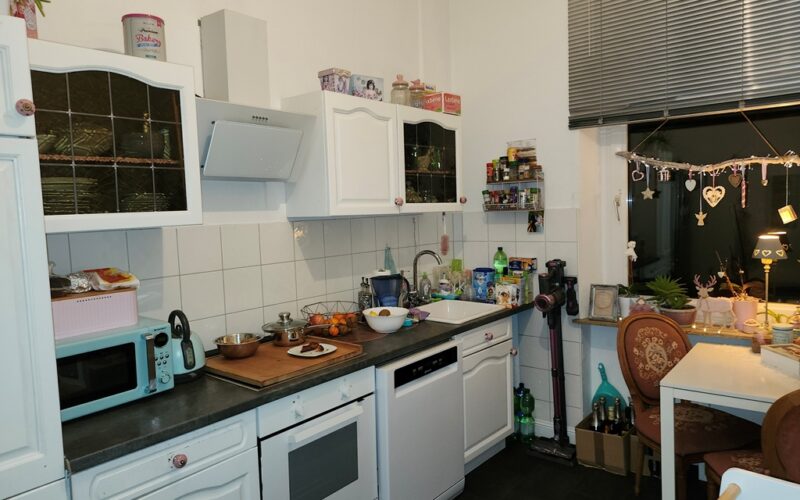 Die Küche im Erdgeschoß mit weißen Einbaumöbeln. Die Wände sind weiß. Das Fenster hat ein Rollo.